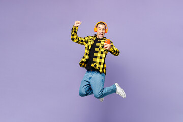 Full body young middle eastern man wear yellow shirt casual clothes jump high listen music in headphones use mobile cell phone do winner gesture isolated on plain purple background. Lifestyle concept.