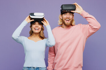 Young fun couple two friends family man woman wear pink blue casual clothes together watching in vr headset pc gadget take off goggles isolated on pastel plain light purple background studio portrait