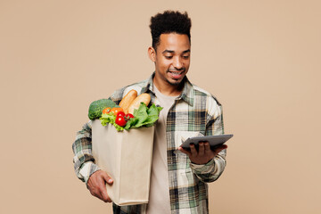 Young smiling fun man wear grey shirt hold paper bag for takeaway mock up with food products use calculator isolated on plain pastel light beige background. Delivery service from shop or restaurant.