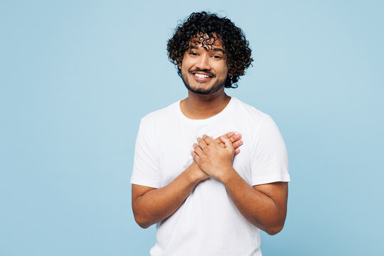 Young smiling grateful happy Indian man wear white t-shirt casual clothes put folded hands on heart look camera isolated on plain pastel light blue cyan background studio portrait. Lifestyle concept.