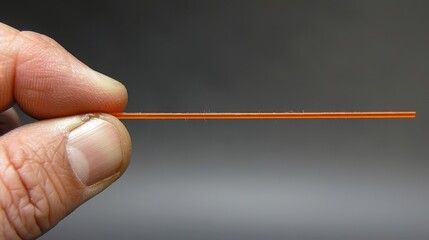   A hand holding a small orange toothpick in tight focus against a gray backdrop - obrazy, fototapety, plakaty