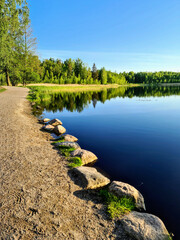 Lakeside Serenity: "A peaceful path runs alongside a tranquil lake, reflecting a clear blue sky, bordered by a line of rocks and the budding greenery of spring
