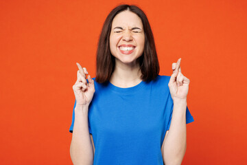 Young smiling happy woman wear blue t-shirt casual clothes waiting for special moment, keeping fingers crossed, making wish isolated on plain red orange background studio portrait. Lifestyle concept. - obrazy, fototapety, plakaty
