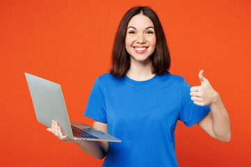 Young smiling satisfied happy IT woman she wear blue t-shirt casual clothes hold use work on laptop pc computer show thumb up isolated on plain red orange background studio portrait Lifestyle concept