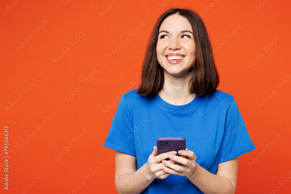 Canvas Prints Young fun surprised happy woman she wear blue t-shirt casual clothes hold in hand use mobile cell phone look aside on area isolated on plain red orange background studio portrait. Lifestyle concept.