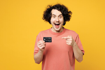 Young excited satisfied happy man wear pink t-shirt casual clothes hold in hand point index finger on mock up of credit bank card isolated on plain yellow orange background studio. Lifestyle concept.