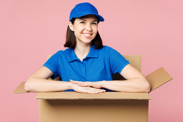 Professional delivery girl employee woman wearing blue cap t-shirt uniform workwear work as dealer courier, posing in big cardboard box look camera isolated on plain pink background. Service concept.