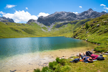 Couple (unrecognizable people) relaxing on the shore of  beautiful La Plagne lake in the French Alps. Other hikers walking up the hill. Peisey valley, Savoie, France. Summer active holidays in nature.
