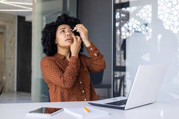 Focused young woman using eye drops at her desk in a bright, contemporary office space. She looks...