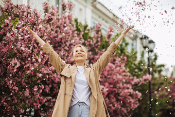 Happy smiling young woman throws up pink petals. Portrait against the backdrop of spring blossoms