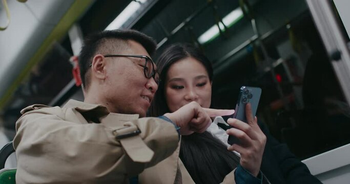 Friends sitting close inside moving bus or train at night. Evening trip. Going back home from work. Watching videos on smartphone. Man pointing on phone screen for friend. Discussing video or image.
