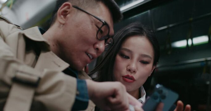 Cute Asian couple traveling together in train or bus. Evening outside window. Man explaining something to girlfriend while typing on phone screen. Woman looking at boyfriend with love.
