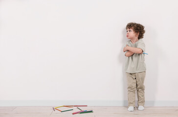Upset little boy with pencil near white wall indoors. Space for text