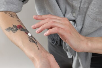 Tattooed woman applying cream onto her hand on light background, closeup