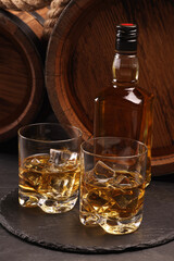 Whiskey with ice cubes in glasses, bottle and wooden barrels on black table