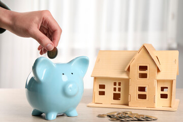 House model and money on wooden table. Woman putting coin into piggy bank indoors, closeup
