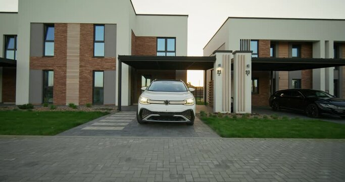 A man drives away from a stylish cottage in a white electric car. A sunny day