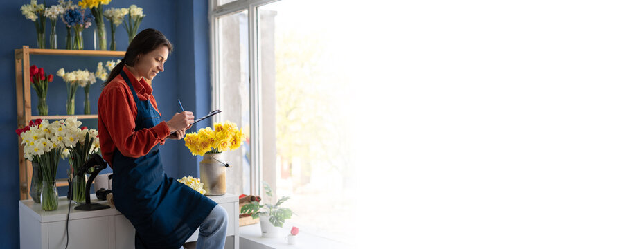 Attractive latin women florist in apron smiling confident writing on document at flower shop looking on the window. Copy space