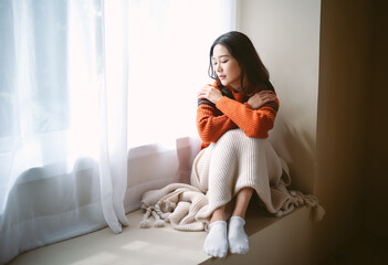 Pensive young Asian woman in warm knitted clothes sitting on windowsill in room. Negative human...