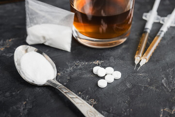 Narcotic substances in spoon, dope powder in transparent plastic bag, syringes with drugs dose, white pills and glass of alcohol drink on dark textured background. Concept of addiction and bad habits
