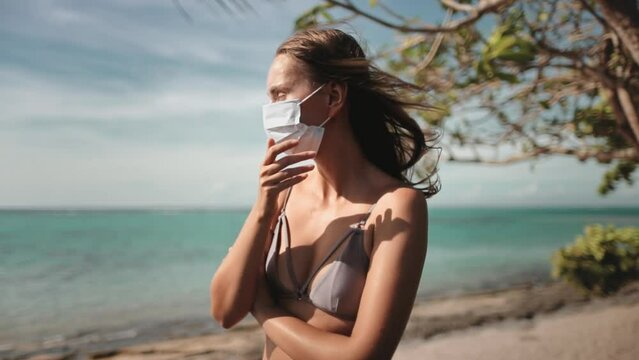 Female in COVID-19 medical mask stands alone on a sunny beach. Girl in mask got sick coughs while have holiday vacation on tropical island. Coronavirus prevention concept. Covid pandemic. Slow motion