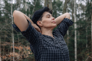 Naklejka premium Happy woman against the backdrop of the forest with her hands behind her head and her eyes closed.