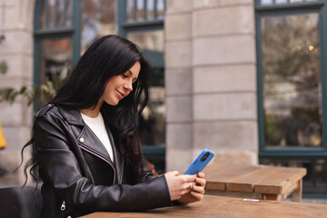 Young beautiful woman holding mobile and looking at smartphone while sitting at cafeteria. Happy girl using mobile phone, read news, shopping and look flirting. Woman smiling, sitting in cafe outdoor.