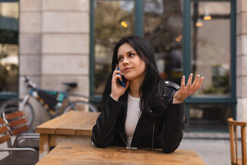 Confused puzzled brunette woman in casual clothes, sits in cafe, have call conversation smartphone,...