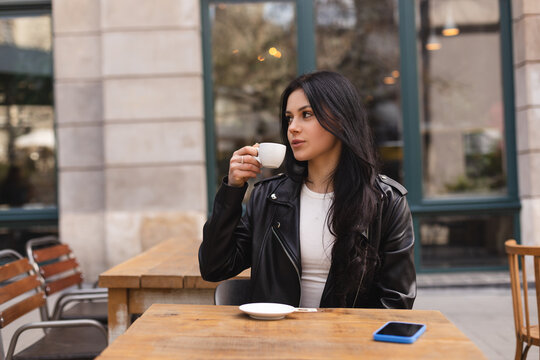 Thoughtful brunette teen woman drinking coffee in cafe outdoor, lifestyle, coffee, communication and local business, unaltered. Sensual woman look at side. Girl wear leather jacker and look pretty.