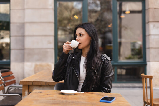 Thoughtful brunette teen woman drinking coffee in cafe outdoor, lifestyle, coffee, communication and local business, unaltered. Sensual woman look at side. Girl wear leather jacker and look pretty.