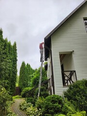 Man on ladder diligently repairing roof of charming white wooden house with determination and hard work under rainy sky, showcasing his craftsmanship in home maintenance.