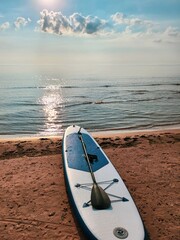 Blue and white paddleboard on sandy beach by calm lake under cloudy sky with sunbeams in morning....