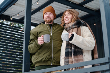 Low angle view medium shot of young Caucasian man and woman in love holding cups with hot drink standing on porch on winter day