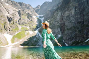 Beautiful woman in summer dress enjoys the view of the mountain lake in sunny weather. Scenery of the majestic mountains. Active lifestyle.