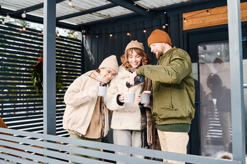 Modern family of three standing on porch on cold winter day, man pouring hot tea from vacuum flask into mugs, copy space