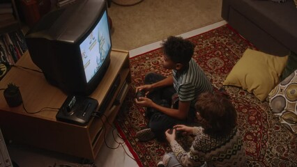 Top view of diverse boys sitting on carpeted floor in living room and playing old-school arcade video game on tube TV using gaming consoles while hanging out together after school, atmospheric 90s