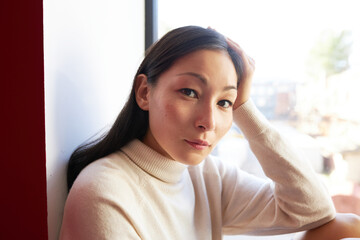 Closeup portrait of gentle charming asian female in sweater sitting on windowsill, having rest...