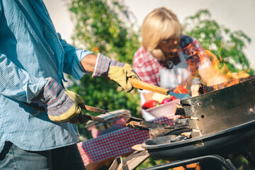 Man wearing protective gloves and holding barbecue tools, while senior woman preparing a food in...