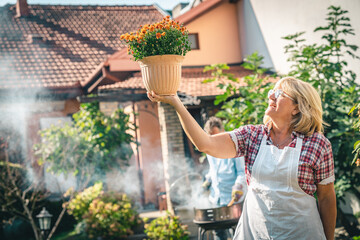 Cheerful senior woman holding a flower pot and admiring her lush plant. Older couple having a...