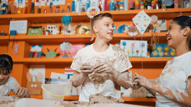Group of happy diverse children playing clay while wearing muddy shirt at art lesson. Cute highschool girl playing with dough while putting clay on happy boy shirt at pottery workshop. Edification.