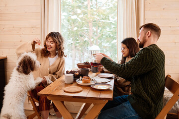 Modern Caucasian family with dog sitting at wooden table in country house enjoying tea party on winter day, copy space