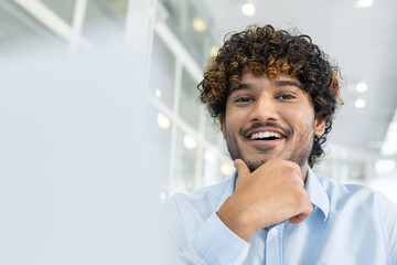 A cheerful young man with curly hair, wearing a light blue shirt, poses with a confident smile in a well-lit modern office setting, exuding positivity and professional confidence.