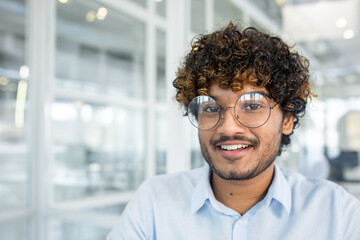 A portrait of a cheerful young man wearing eyeglasses, smiling at the camera in a bright, contemporary office environment. The image exudes positivity and confidence.