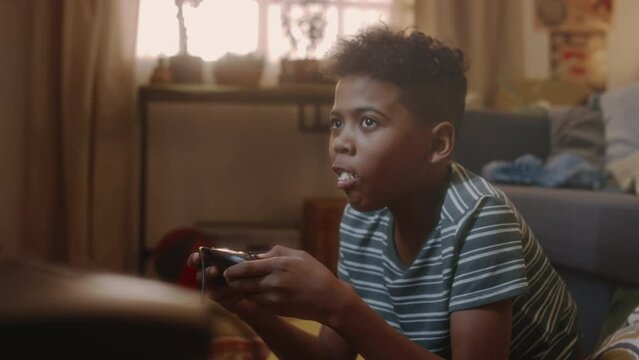 Tilt handheld footage of African American gen Y kid eating handful of popcorn while playing console video game in front of TV sitting on floor in retro-styled living room, atmosphere of 90s