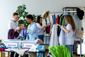 Group of volunteers, young women and man, sorting clothes in charitable foundation for charity...