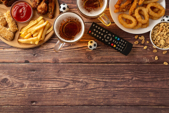 An inviting display of game day snacks including fries, chicken wings, onion rings, and beer, arranged neatly next to a remote control and mini soccer balls
