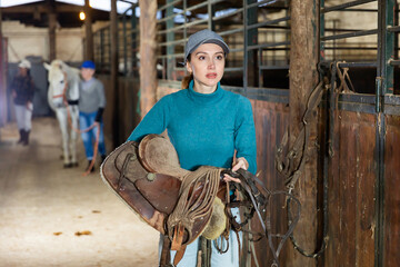 European woman horse breeder carrying leather saddle in horse barn.