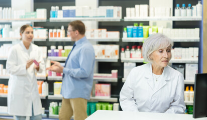 Senior woman pharmacist in pharmacy works with computer, searches among nomenclature of drugs, specifies price and availability of medicine - obrazy, fototapety, plakaty