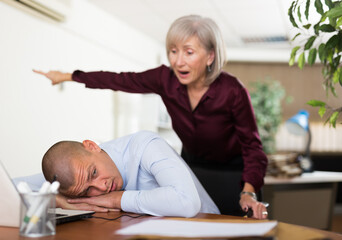 Man office worker sleeping on table during work day. His chief senior woman standing next to him...