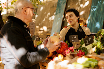 At farmers market, woman vendor displaying fresh organic squash to old man. Small business female owner standing behind a fruit and vegetable stand, offering customers wholesome locally grown produce.